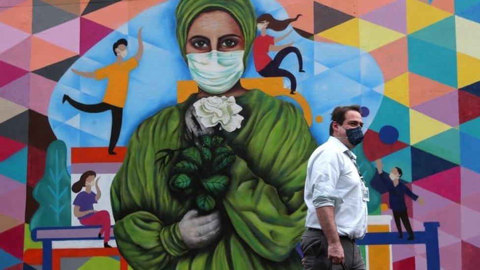 Man in front of a mural with a drawing of a woman with a mask.