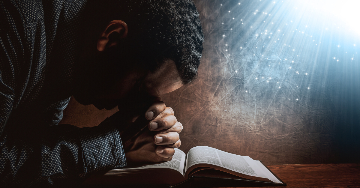 man resting head on praying hands on open bible light streaming above holy spirit