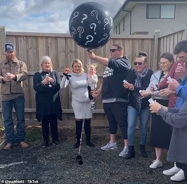 The gender reveal begins with people gathered around, ready to pop a big black balloon
